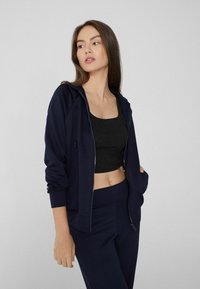 Sudadera navy con cremallera y ajuste relajado, puños acanalados y bolsillos tipo canguro, combinada con una camiseta de corte ajustado negra. Textura de tela suave.