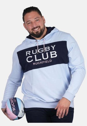 Homme souriant, portant un sweat à capuche bleu clair avec l'inscription "Rugby Club Ruckfield", tenant un ballon de rugby multicolore dans sa main gauche.