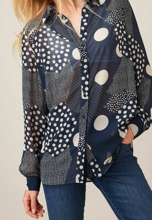 Woman wearing a navy blue sheer blouse with mixed polka dot patterns paired with dark blue jeans, hand in pocket.