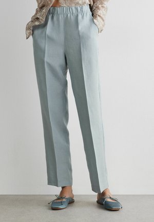 Lyseblå højtaljede bukser med frontplisseringer, båret med turkisblå flade sko med sløjfe, kombineret med en beige mønstret bluse, der er stoppet ind.