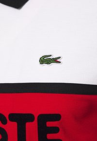 Pikepaita valkoisena, jossa on punaiset ja mustat raidat, rintalogona vihreä brodeerattu krokotiili. Voimakkaasti teksturoitu kangas.