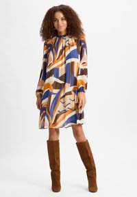 Mehrfarbiges kurzes Kleid mit langen Ärmeln, das ein Muster aus blauen, orangefarbenen, braunen und cremfarbenen Formen aufweist. Braune Stiefel bis zum Knie vervollständigen den Look.