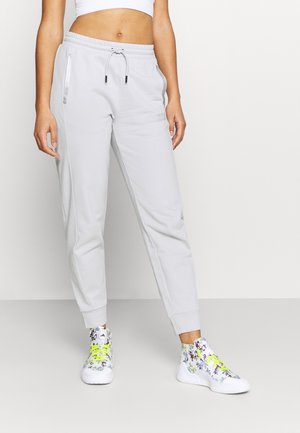 Femme portant un pantalon jogger blanc taille haute et des baskets montantes blanches à imprimé floral avec des lacets jaune fluo, debout devant un fond uni.