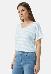 Jasnoniebieska koszulka z dekoltem w serek, zdobiona poziomymi paskami i wzorem ptaków. Krótkie rękawy, luźny krój, wykonana z miękkiego materiału. Noszona z niebieskimi dżinsami.