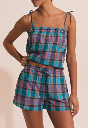 Kobieta ubrana w turkusowy, fioletowy i różowy kraciasty top bez rękawów z wiązanymi ramiączkami i dopasowane krótkie spodenki ze sznurkiem, stojąca na jednolitym tle.
