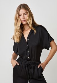 Top nero a maniche corte in tessuto liscio con colletto e chiusura a bottoni. Abbinato a pantaloni neri a vita elasticizzata e vestibilità rilassata.