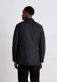Veste noire en tissu texturé avec une coupe décontractée. Comprend une fente au dos, des boutons pression et une petite étiquette au col.