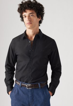 Black long-sleeve shirt with buttons, featuring a pointed collar. Worn with a woven brown leather belt and blue casual trousers.