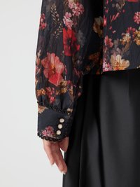 Bluse med blomstermønster i svart gjennomsiktig stoff, prydet med røde og rosa blomster. Ermet har svart blondertrim og tre dekorative knapper.