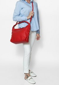 Borsa a tracolla in pelle rossa con manico intrecciato, forma rettangolare, texture liscia e tracolla regolabile. Abbinata a una camicia azzurro chiaro e pantaloni bianchi.