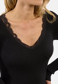 Donna che indossa un maglione a coste nero con scollatura a V e delicati bordi in pizzo nero lungo il colletto, che mostra parte del suo collo e della spalla.