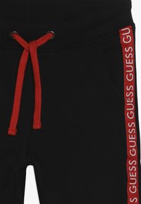 Černé tepláky s měkkou strukturou, vybavené červeným šňůrkem a bočním pruhem s opakujícím se bílým logem "GUESS".