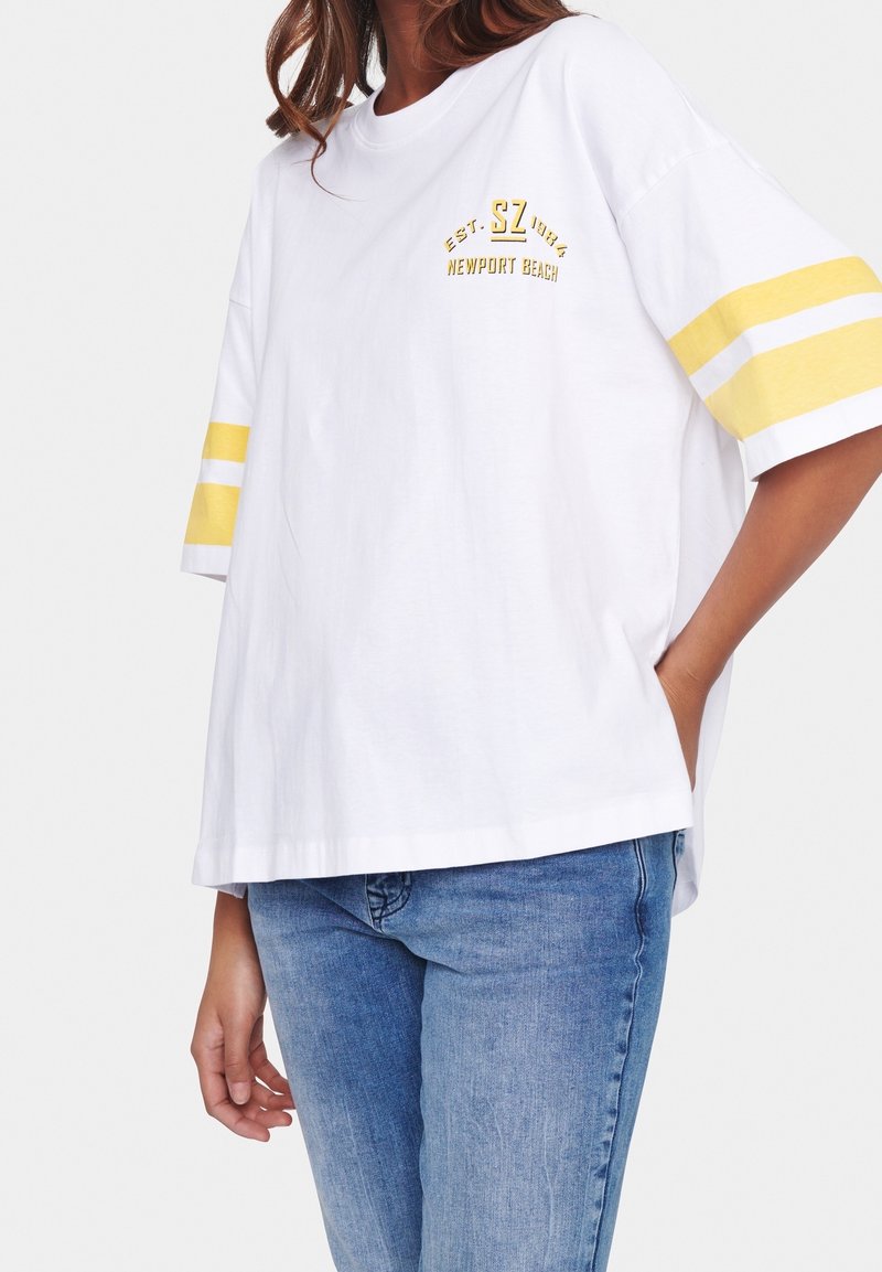 Žena oblečená v volné bílé tričko se žlutými pruhy na rukávech a textem "Newport Beach", v kombinaci s modrými džínami, na bílém pozadí.