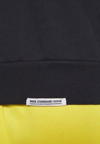 Tela negra con una etiqueta blanca de Nike Standard Issue encima de una tela de malla amarilla, mostrando el texto "El estilo clásico se une al rendimiento moderno."