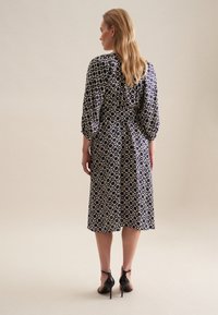 Granatowa sukienka w wzory z rękawami o długości trzech czwartych, wąską talią i do wysokości łydki, zestawiona z czarnymi szpilkami.
