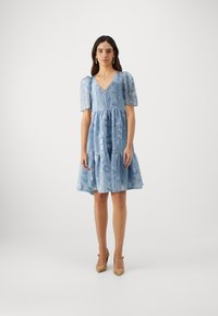 Robe bleu clair, longue jusqu'aux genoux, avec des manches courtes bouffantes, un décolleté en V et des détails en texture florale. Présente une jupe à volants et un tissu doux.