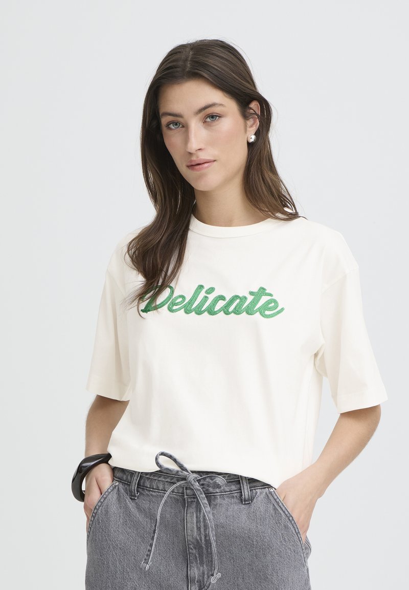 T-shirt en coton couleur crème avec le mot « Delicate » en broderie verte. Manches courtes, coupe décontractée, portée avec un short en jean gris taille haute.