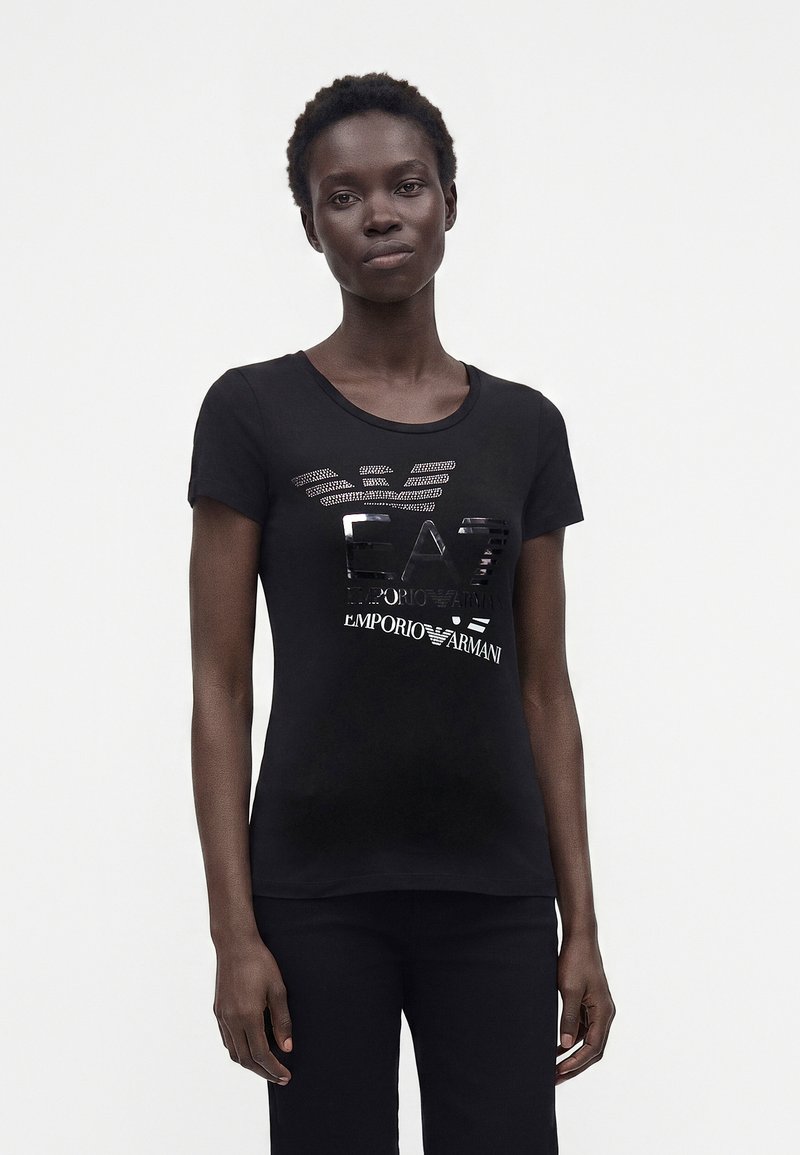 Νεαρή γυναίκα που φοράει μαύρο t-shirt Emporio Armani με γυαλιστερό λογότυπο, στέκεται μπροστά σε ένα απλό ανοιχτό φόντο.