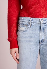 Röd stickad tröja med ribbad textur, matchad med ljusblå slitna denimjeans med fem fickor och metallknappsstängning.