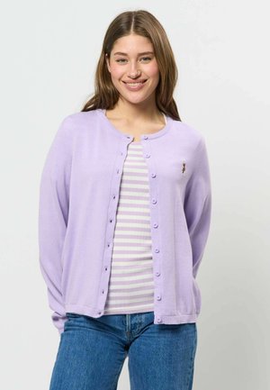 Ung kvinde smiler, iført en lyse-lilla knapcardigan over en hvid og lilla striped top og blå jeans, stående mod en ensfarvet baggrund.