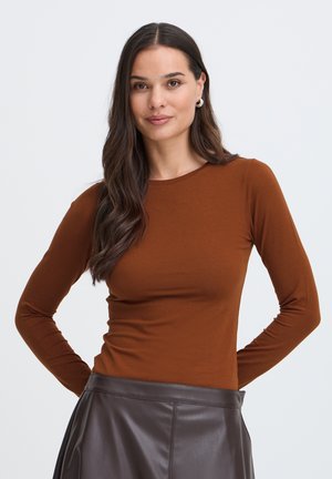 Femme aux longs cheveux foncés porte un haut marron à manches longues et une jupe en cuir marron foncé, debout les mains derrière le dos sur un fond uni.