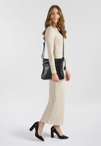 Borsa a tracolla in pelle nera con una texture liscia, tracolla regolabile e tasca frontale con zip. Modella che indossa un lungo abito beige chiaro e tacchi neri.