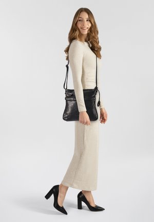 Borsa a tracolla in pelle nera con una texture liscia, tracolla regolabile e tasca frontale con zip. Modella che indossa un lungo abito beige chiaro e tacchi neri.