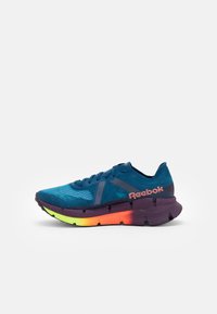 Zapatilla deportiva azul con parte superior de malla, media suela morada en contraste y suela exterior en degradado de naranja a amarillo. Presenta la marca Reebok en el costado.