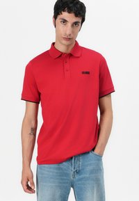 Κόκκινο πόλο μπλουζάκι με κανονική εφαρμογή, κοντά μανίκια, μαύρες λεπτομέρειες στις μανσέτες και μικρή λογότυπο στο στήθος. Φτιαγμένο από αναπνέον ύφασμα.