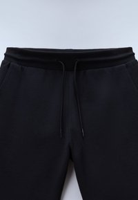 Musta collegehousut, joissa on resorivyötärö, säädettävä kiristysnyöri ja sivutaskut. Pehmeä kangas, sileä pinta ja yksinkertainen muotoilu.