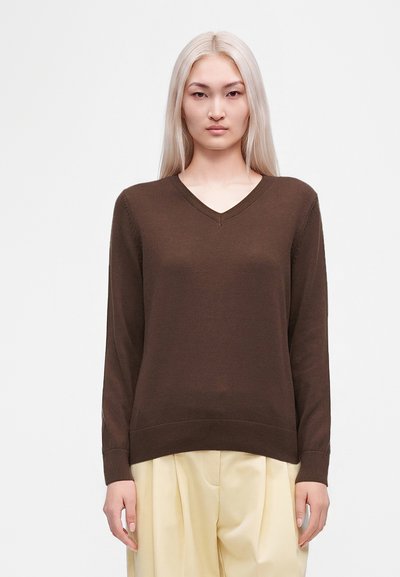 Femme aux longs cheveux blonds, portant un pull marron à col en V et un pantalon plissé beige clair, debout devant un fond blanc uni.