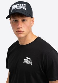 Tricou din bumbac negru cu un logo alb pe piept; șapcă neagră cu "Lonsdale London" brodată în literă albă.