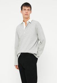 Lichtgrijze polo met lange mouwen en witte kraag en drie knopen, gecombineerd met zwarte broek. Zachte stof met een ontspannen pasvorm.