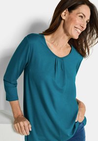 Türkise Bluse mit dreiviertel Ärmeln, einem runden Ausschnitt und Faltendetail vorne, aus einem weichen, glatten Stoff gefertigt.