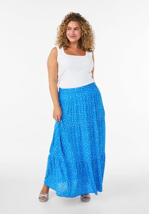 Frau mit lockigem Haar, die ein weißes ärmelloses Top, einen langen blauen Rock mit Blumenmuster und hellblaue Sandalen mit Absatz trägt, steht vor einem weißen Hintergrund.