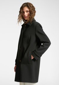 Abrigo negro elaborado en tela texturizada, con cuello, bolsillos laterales y acentos de botones dorados. Largo hasta la rodilla con una silueta recta.