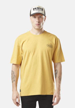 T-shirt en coton jaune à manches courtes avec un col rond et un petit patch logo noir sur la poitrine, associé à une casquette trucker noire et crème.