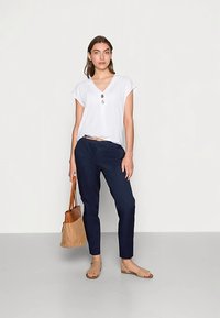 Witte top met korte mouwen en knopen, marineblauwe taps toelopende broek, beige sandalen en een gevlochten mandtas. Model staat tegen een neutrale achtergrond.