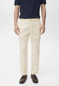 Pantalones de lino beige con cordón en la cintura, corte recto y bolsillos laterales. Llevados con una camisa texturizada azul marino y zapatos marrones.