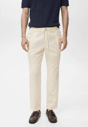 Pantalones de lino beige con cordón en la cintura, corte recto y bolsillos laterales. Llevados con una camisa texturizada azul marino y zapatos marrones.