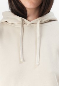 Hoodie beige avec une capuche à cordon, texture douce et ourlet côtelé. Le tissu est lisse et légèrement extensible, adapté pour un usage décontracté.