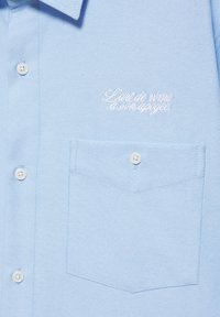 Chemise bleu clair avec des boutons blancs et une poche poitrine, brodée d'un texte français blanc en cursive sur le haut à gauche.