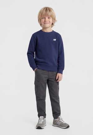 Mladý chlapec stojí, oblečený v námorníckej modrej mikine, tmavých maskáčových nohaviciach a sivých teniskách New Balance, usmieva sa s rukami v vreckách.