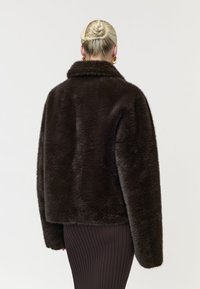 Femme blonde avec un chignon tressé portant une veste en fausse fourrure marron foncé et une jupe côtelée, vue de dos sur un fond uni.