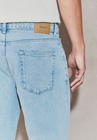 Light blue denim jeans with a visible back pocket and a tan leather patch featuring the text "DENIM SUPPLY." Smooth texture and classic fit.