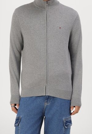 Personne portant un pull gris à fermeture éclair avec un petit logo sur la poitrine et un jean cargo bleu, debout devant un fond uni.