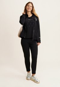 Felpa nera con zip, t-shirt nera con grafica e joggers neri a taglio stretto. Sneakers argentate. Completa l'outfit una borsa a spalla di colore chiaro.