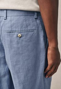 Pantalones de lino azul claro con un único bolsillo trasero con un botón crema, delantero plano y textura sutil. Vista lateral que muestra el ajuste.
