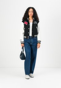 Giacca varsity bianca e nera con toppe ricamate, maniche lunghe e polsini a righe, abbinata a jeans blu a vita alta e scarpe bianche.