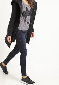 Black long coat over a gray t-shirt with a lace-patterned Minnie Mouse design, dark skinny jeans, and black sneakers with white soles.